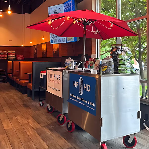 A Hi Five Hotdogs cart with a red umbrella inside the restaurant, featuring booths and large windows.